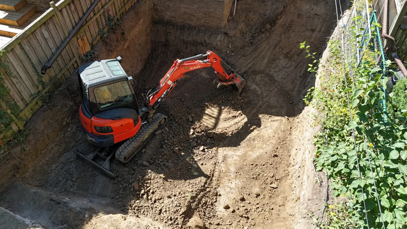 Orange Pinemar compact excavator working in a tight side-yard excavation beside a brick building