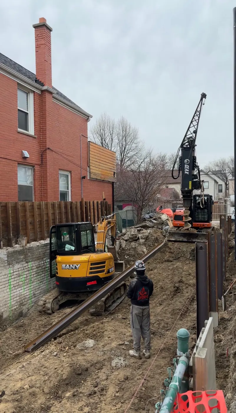 Excavator and drilling equipment working beside a residential shoring installation