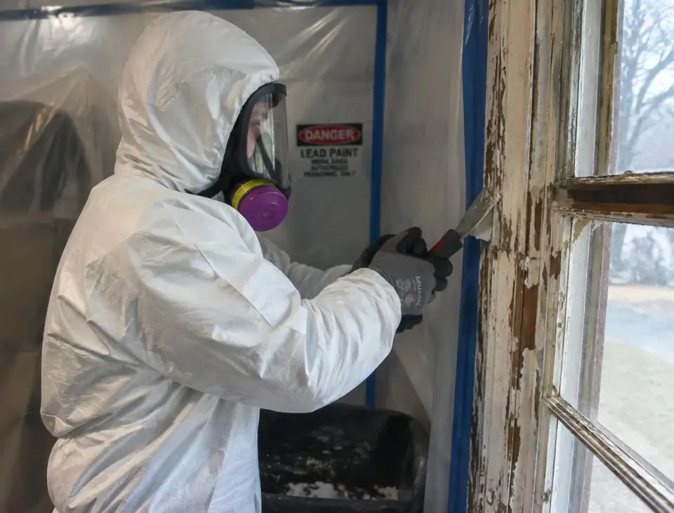 Worker in protective suit scraping old lead paint from a window frame inside containment